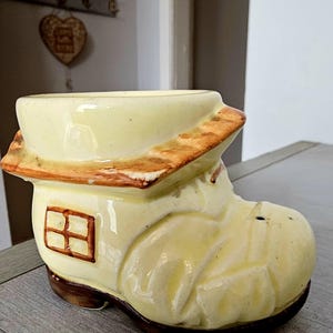 May include: A pale yellow ceramic planter shaped like a shoe with a house design. The shoe features a brown roof-like rim, a small brown window on the side, and a dark brown sole. The surface has a glossy finish with subtle molded details.