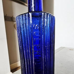 Könnte beinhalten: Eine kobaltblaue Vintage-Glasflasche mit rechteckigem Körper und vertikal gerippter Textur. Das Wort SULPHOLINE ist vertikal in Großbuchstaben auf der Vorderseite eingeprägt. Die Flasche hat einen schmalen Hals und einen abgerundeten Rand.