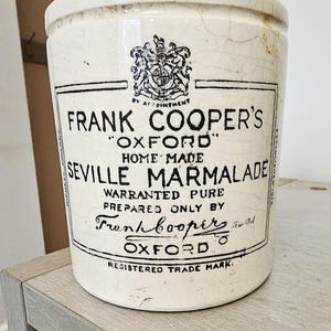 May include: A cream-coloured ceramic crock with a crackled glaze finish. The front features black printed text reading Frank Cooper's Oxford Home Made Seville Marmalade, along with a crest and the words Registered Trade Mark.