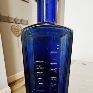 Op de afbeelding: Een rechthoekige kobaltblauwe glazen fles met een smalle hals en een dikke rand. De voorzijde van de fles is voorzien van reliëftekst met de tekst LILY BALM en (REGD) in hoofdletters. Het glas heeft een gladde, glanzende afwerking en diepblauwe kleur.