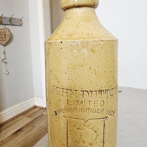Könnte beinhalten: Eine hohe, zylindrische Ingwerbierflasche aus beigem Steingut mit abgerundetem Oberteil. Die Flasche zeigt den geprägten Text JOHN DUTTON LIMITED SUPERIOR GINGER BEER, ein zentrales Schild-Logo mit dem Buchstaben D und MANCHESTER am Boden.