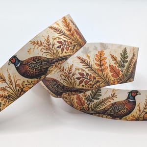 Op de afbeelding: Een rol crèmekleurig lint met een herhalend patroon van fazanten en herfstkleurig gebladerte, waaronder oranje, bruine en groene bladeren. Het ontwerp toont gedetailleerde illustraties van vogels met blauwe koppen en gevederde patronen.