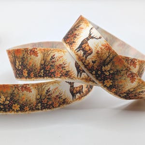 Könnte beinhalten: Ein Stoffband mit einem sich wiederholenden herbstlichen Waldmotiv mit braunen Hirschen, orangefarbenen und gelben Ahornblättern sowie dunklen Beeren auf cremefarbenem Grund. Das Band ist aufgerollt und zeigt den detaillierten Waldaufdruck.