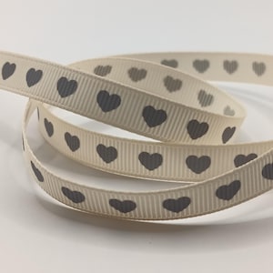 May include: Cream-coloured ribbon featuring a repeating pattern of grey hearts. The ribbon has a tightly woven, ribbed texture, with evenly spaced hearts. The ribbon is curled and looped, creating a decorative effect, suitable for crafts.