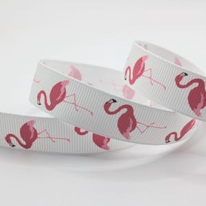 May include: White ribbon featuring a repeating pattern of pink flamingos. The flamingos are illustrated in a simple, cartoon style. The ribbon is curled, suggesting it's made of a durable material, ideal for crafts or gift wrapping.