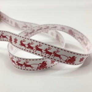 May include: White ribbon featuring a repeating pattern of red reindeer and Santa's sleigh, with snowflakes. The ribbon has a stitched border design. The ribbon is curled and looped, showing the pattern on both sides.