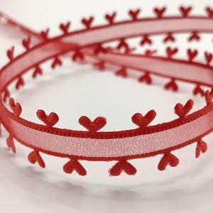 May include: Red ribbon featuring embroidered heart-shaped details. The ribbon has a semi-transparent quality, with a woven texture in the middle. Heart motifs are evenly spaced along the edges, forming a decorative trim. Suitable for crafts and embellishments.