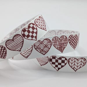 May include: White ribbon decorated with maroon heart patterns. The hearts have various designs, such as stripes and checks. This ribbon is suitable for crafts or gift wrapping. The ribbon's width is approximately 2.5 cm.