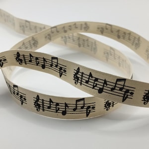 May include: A coiled length of off-white fabric ribbon featuring a repeating black print of musical notation, including treble clefs, staff lines, and various musical notes arranged in a continuous pattern along the length of the woven material.