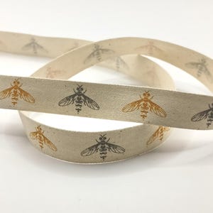 May include: Beige ribbon featuring a repeating pattern of bees in brown and grey. The ribbon is curled, suggesting a natural fabric. The bees are detailed with wings and body segments, creating a decorative effect.