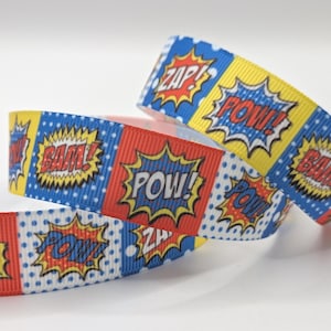 Pode incluir: Um rolo de fita gorgurão com um padrão de banda desenhada com painéis alternados em vermelho, amarelo e azul. O design inclui bolinhas brancas e balões de fala estilo banda desenhada com as palavras ZAP, POW e BAM em letras negrito.