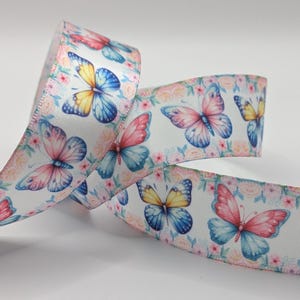 May include: White ribbon featuring a repeating pattern of butterflies and small pink flowers. The butterflies have blue, yellow, and pink wings. The ribbon is curled, displaying the pattern on both sides.