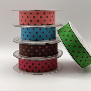 May include: A selection of decorative ribbon spools in shades of red, blue, brown, and green. Each spool features a repeating star pattern in contrasting colours. The ribbons are stacked on clear plastic spools.