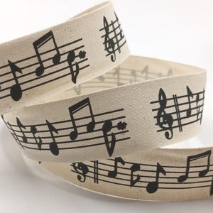 May include: Cream-coloured ribbon with a repeating pattern of black musical notes and treble clef symbols. The ribbon is made of woven fabric, suitable for crafting or gift wrapping. The musical notes are printed in a clear, bold font.