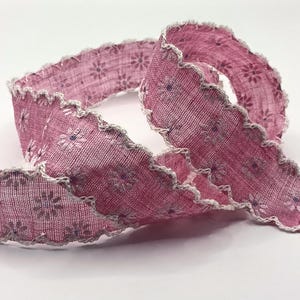 May include: A length of pink ribbon with a floral pattern and a scalloped edge. The ribbon is a dusty rose colour with small white and purple flowers. The edges are trimmed with a white, textured trim. The ribbon is curled and folded.
