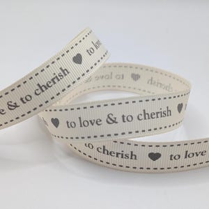 May include: Cream-coloured ribbon with the text "to love & to cherish" and heart symbols. The ribbon has a dotted border and is coiled in a loose arrangement. The text and hearts are in a dark grey colour.