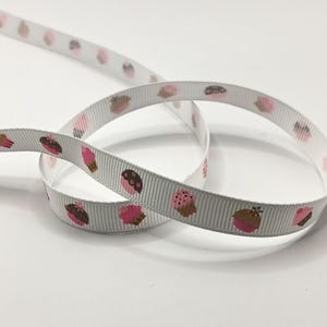 May include: White ribbon featuring a repeating pattern of cupcakes in pink, brown, and beige. The ribbon is curled on a white surface. The cupcakes have different designs, including sprinkles and icing.