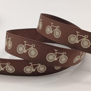 May include: A length of brown ribbon featuring a repeating design of bicycles in a lighter tone. The ribbon is folded, displaying the bicycle pattern. The bicycles have detailed spokes and frames, ideal for crafting or gift wrapping.