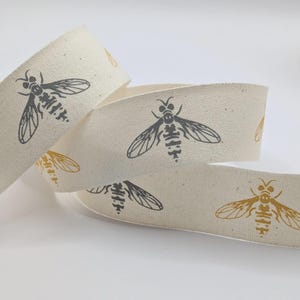 May include: Cream-coloured ribbon featuring a repeating pattern of stylised bees in grey and gold. The woven fabric ribbon is curled in a loose spiral, displaying the bee design. Suitable for crafts and gift wrapping.