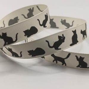 May include: Cream-coloured ribbon featuring a repeating pattern of black cat silhouettes in different poses. The ribbon has a ribbed texture and is curled in a loose spiral, displaying the cat design. Suitable for crafts and gift wrapping.