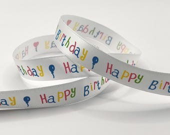 Könnte beinhalten: Ein weißes Satinband mit einem sich wiederholenden Muster aus dem Text Happy Birthday in bunten Buchstaben, darunter Blau, Rot, Gelb und Grün, durchsetzt mit kleinen blauen Ballonsymbolen, die entlang des Stoffes aufgedruckt sind.