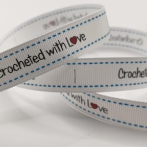 May include: White ribbon with the words "crocheted with Love" printed in black, with a red heart. The edges are detailed with blue dashed lines. The ribbon is curled and layered.