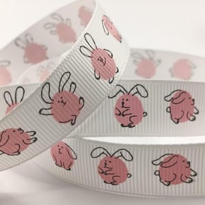 May include: White ribbon featuring a repeating pattern of pink bunny illustrations. Each bunny is drawn in a cartoon style with black outlines. The ribbon is folded over itself, showing the pattern at different angles.