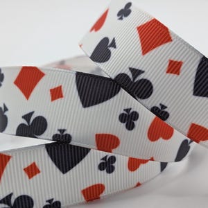 May include: White ribbon patterned with black and red playing card suits: spades, hearts, diamonds, and clubs. The ribbon is suitable for crafts or gift wrapping. The ribbon's width is approximately 2.5 cm.