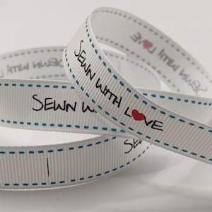 May include: White ribbon featuring the text "SEWN WITH LOVE" in black, with a red heart. The ribbon has a blue dotted border. The words "SEWN WITH LOVE" are repeated on the ribbon.