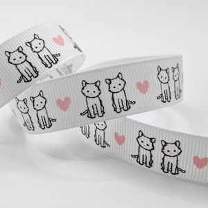 May include: White ribbon featuring a repeating pattern of black cat illustrations and pink hearts. The ribbon is curled and layered, displaying the design. Suitable for crafts, gift wrapping, or adding a decorative touch.