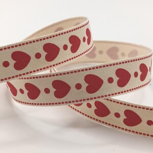 May include: Cream-coloured ribbon featuring a repeating pattern of red hearts and dots. The edges are lined with a dotted red border. The ribbon is curled and layered, displaying the design.