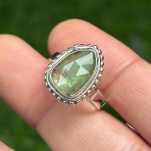 May include: A silver ring featuring a pear-shaped, faceted green gemstone. The gemstone is set in a bezel setting and surrounded by a beaded border. The ring's band is a simple, polished silver.