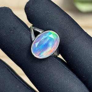 May include: A silver ring featuring a large, oval-cut gemstone with a rainbow iridescence. The gemstone displays a spectrum of colors, including blue, green, and pink. The ring's band has a simple, geometric design.