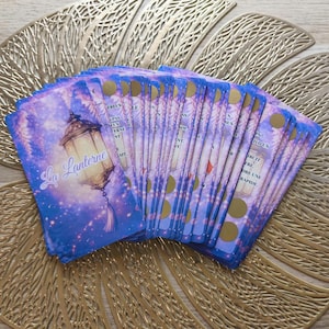 Peut inclure: Un jeu de cartes oracle avec un design céleste. Les cartes présentent un fond étoilé violet et bleu avec une illustration de lanterne et le texte "La Lanterne". Chaque carte a des accents dorés et du texte.
