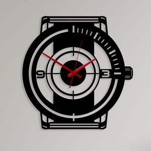May include: Black wall clock in the shape of a wristwatch. The clock features a circular face with red hands and a black frame. The numbers 3 and 9 are visible. The clock is mounted on a white wall.