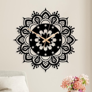 May include: A black, laser-cut mandala wall clock with wooden clock hands. The clock is mounted on a white wall above a gray armchair. A small table with a vase of flowers and an alarm clock is in front of the chair.