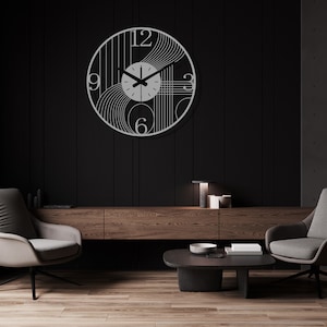 May include: A modern, minimalist wall clock with a silver metal frame and black hands. The clock face features a geometric design with lines and curves. The clock is hanging on a black wall in a living room setting with two armchairs and a coffee table.