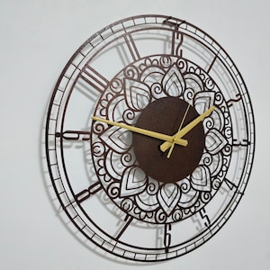 Pode incluir: Um relógio de parede grande, redondo, de metal marrom com marcas de números romanos e um design de mandala decorativo. O relógio tem ponteiros dourados.