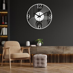 May include: A white clock with black lines and numbers on a black wall. The clock is hanging above a brown wooden shelf with a tan armchair and a gray ottoman in front of it.