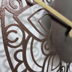 May include: Close-up of a copper-colored metal wall clock with a decorative, intricate, cut-out design. The clock has a gold-colored hand.