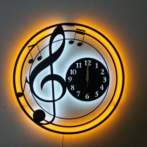 Puede incluir: Un reloj de pared de metal negro con un diseño de clave de sol y una esfera blanca. El reloj tiene una luz LED amarilla alrededor del borde.