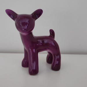 Puede incluir: Una figurita de cerámica de ciervo de color púrpura. El ciervo está de pie sobre sus cuatro patas con la cabeza en alto. La figurita es brillante y tiene un acabado liso.