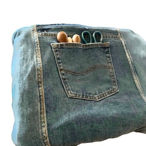 Op de afbeelding: Een blauwe denim tas met een groot vak aan de voorkant. Het vak bevat houten gereedschap en een groene schaar.
