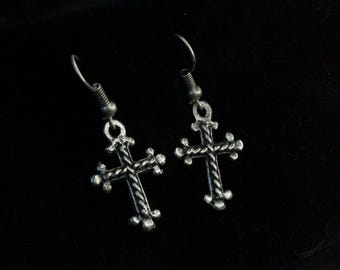 May include: A pair of silver cross earrings. Each earring has a detailed cross design with a textured surface and small decorative elements at the ends of the cross arms. The earrings hang from a simple hook.