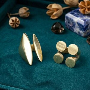 K&ouml;nnte beinhalten: Zwei goldfarbene Ringe auf einer teal-farbenen Oberfl&auml;che. Ein Ring hat eine dreieckige Form mit einem langen, ovalen Akzent. Der andere Ring hat vier kreisf&ouml;rmige Akzente. Dekorative Elemente und ein Edelstein im Hintergrund.