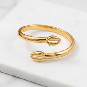 Puede incluir: Una pulsera de puño dorada con un diseño en espiral. La pulsera presenta dos adornos en forma de concha en cada extremo. La joya se exhibe sobre una superficie de mármol blanco.