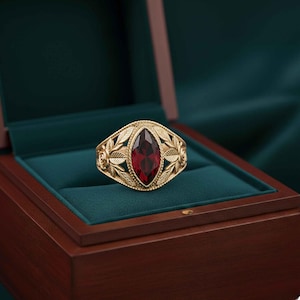 May include: A gold ring with a large, oval, deep red gemstone. The ring has a detailed leaf and floral design. It's displayed in a wooden jewellery box with a green velvet interior. The ring's design is intricate and eye-catching.