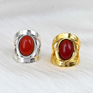 May include: Two rings, one silver and one gold, each featuring a large, oval, red gemstone. The rings have a unique open design around the stone, creating a striking visual contrast. The gemstones are a vibrant red.