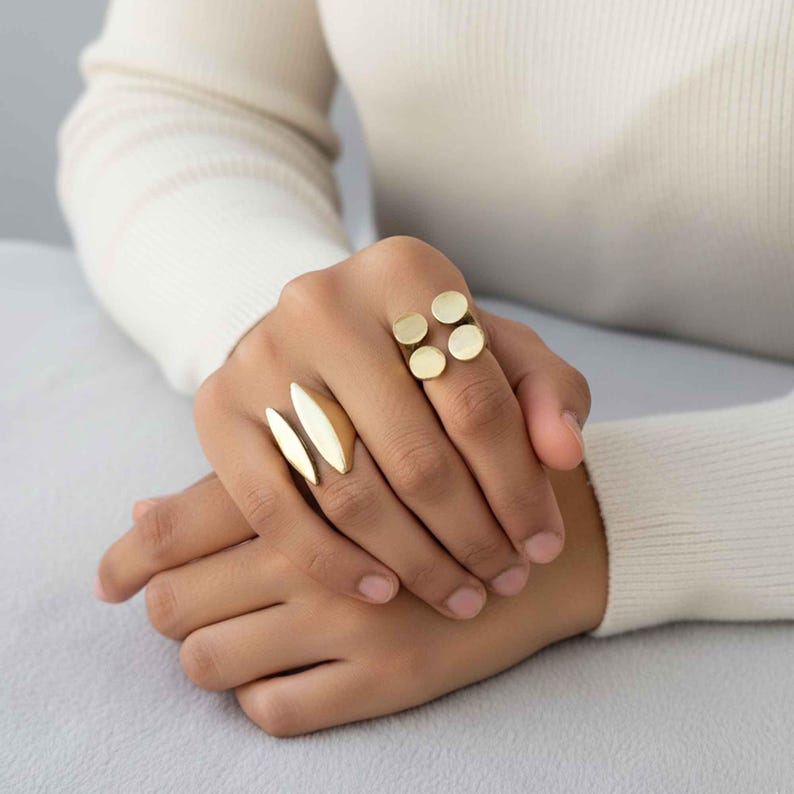 K&ouml;nnte beinhalten: Nahaufnahme einer Hand mit goldfarbenen Ringen. Ein Ring hat zwei l&auml;ngliche, blattf&ouml;rmige Designs, w&auml;hrend ein anderer vier kreisf&ouml;rmige Akzente aufweist. Die Ringe werden an verschiedenen Fingern getragen und zeigen ihre einzigartigen Stile und das metallische Finish.