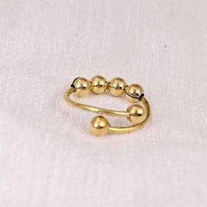 Puede incluir: Un anillo ajustable de color dorado con cinco pequeñas cuentas redondas y un diseño en espiral. El anillo tiene un acabado liso y pulido sobre un fondo blanco. El anillo es una joya.